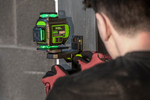 Tradesperson in work gloves fitting a Grettz green beam multi-line laser to a wall bracket, with bright green 360-degree laser lines projected across a grey blockwork wall