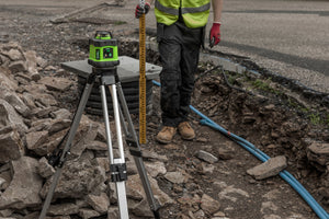 Groundworker in hi-vis holding a levelling staff beside blue pipework and rubble in an open trench, with a Grettz green rotary laser level set up on a tripod behind