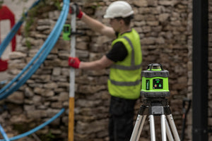 Builder in hi-vis and hard hat checking a reading on a levelling staff against a stone wall, with a Grettz green rotary laser level set up on a tripod in the foreground