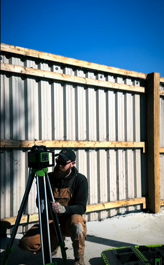 Tradesperson kneeling to set up a Grettz green laser level on a tripod in front of a timber fence and blockwork under a blue sky
