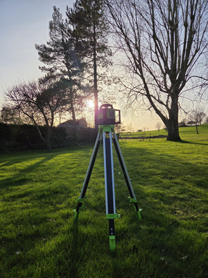 Grettz green rotary laser level set up on a tripod on grass at dusk, sun setting through bare trees behind on an outdoor site